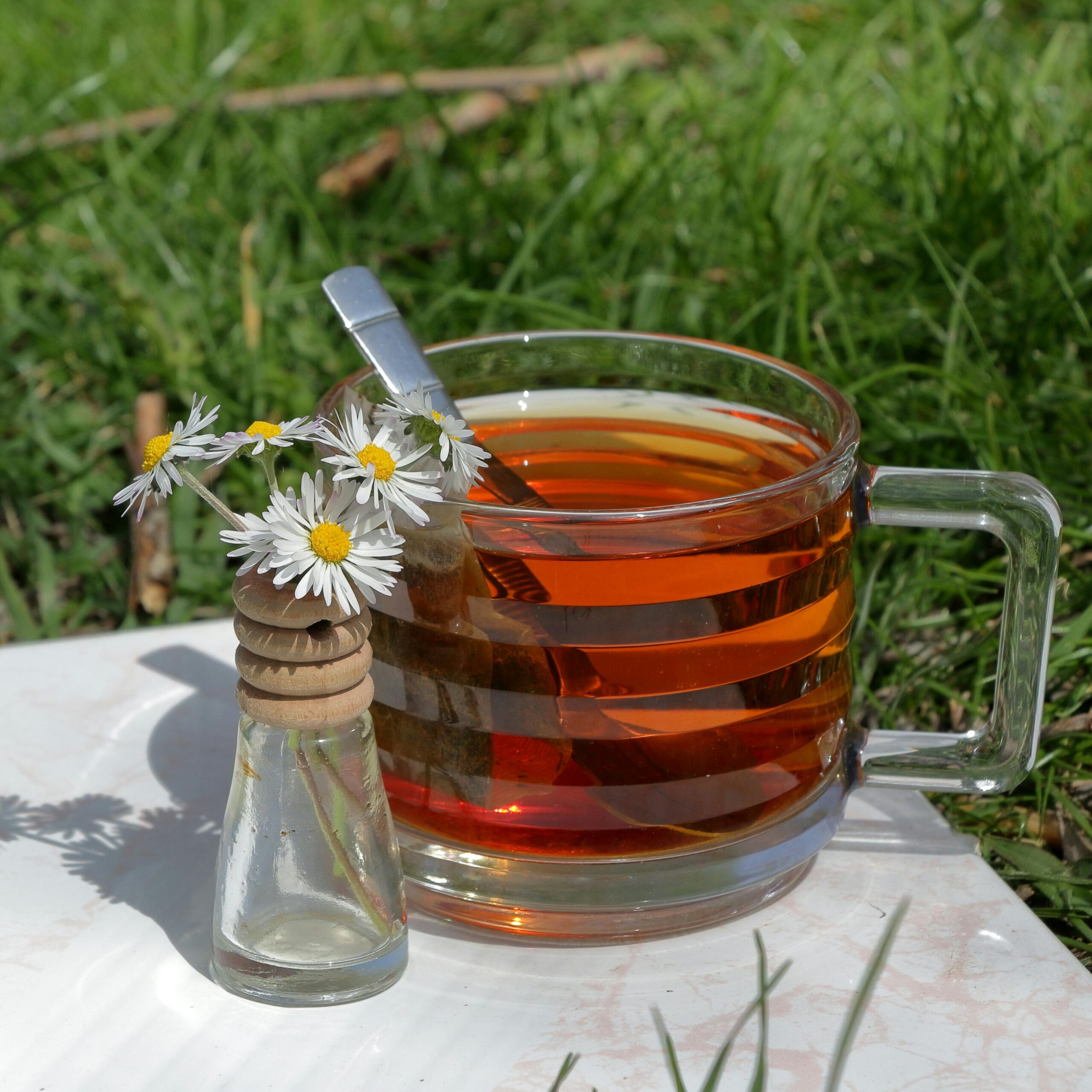 A hot herbal tea served in a glass mug with chamomile flowers in a bottle on grass.